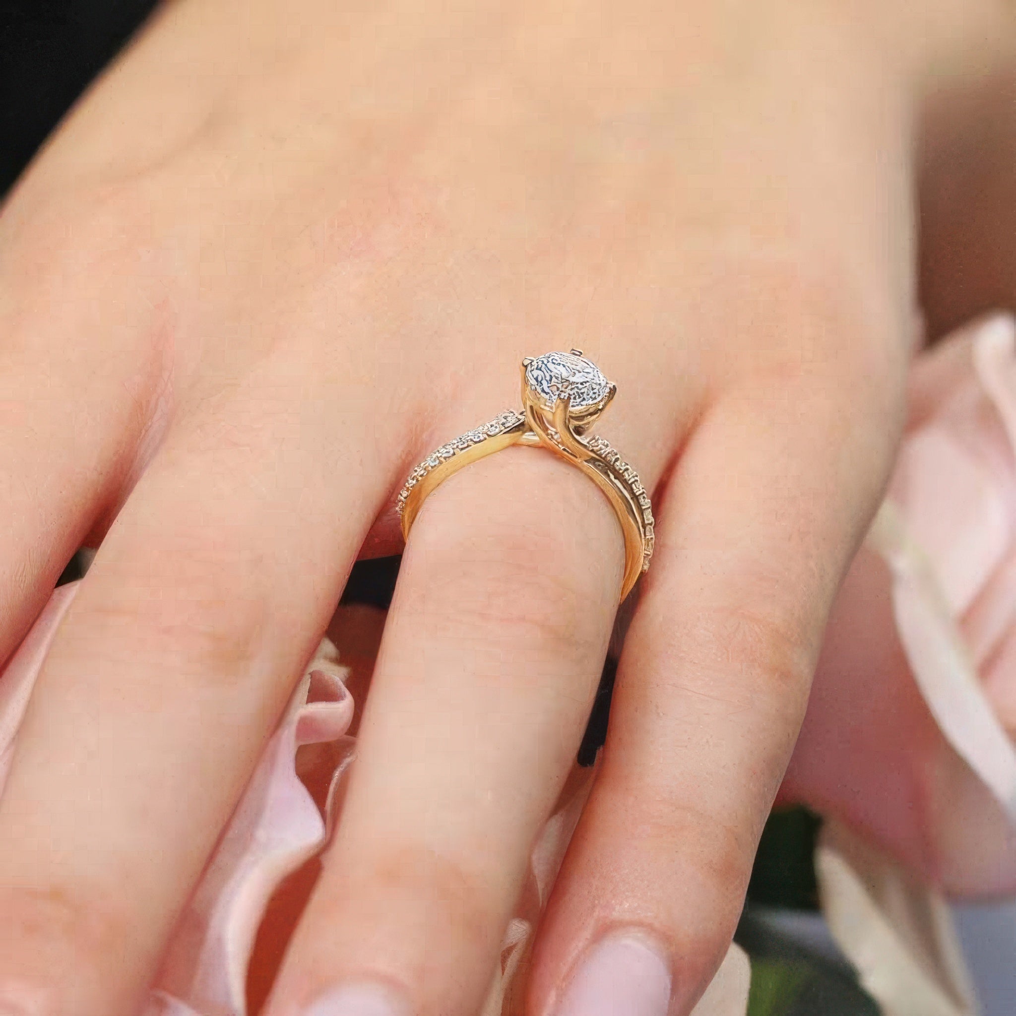 Elegant twisted engagement ring with a round center stone, worn on a hand among soft pink flowers.