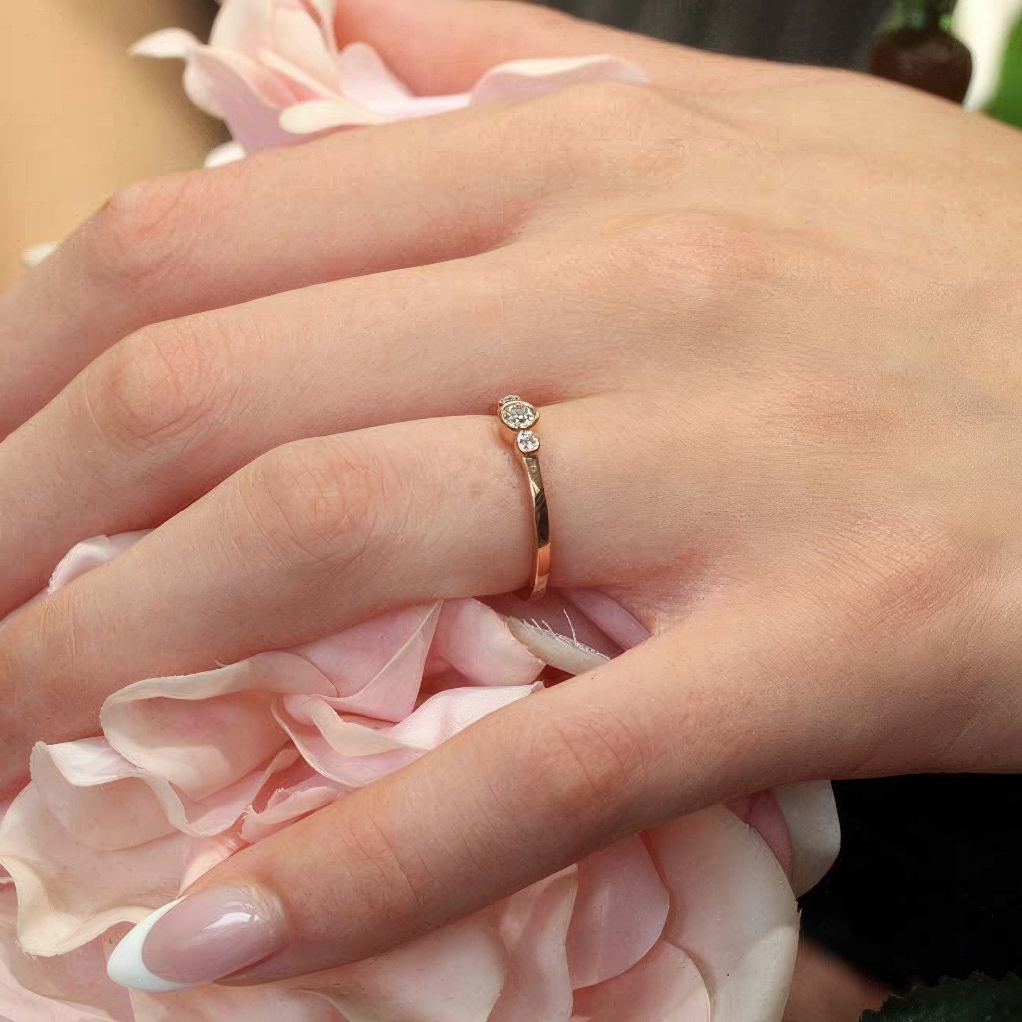 Close-up of a hand wearing a white gold three stone promise ring with diamonds, resting on pink rose petals.