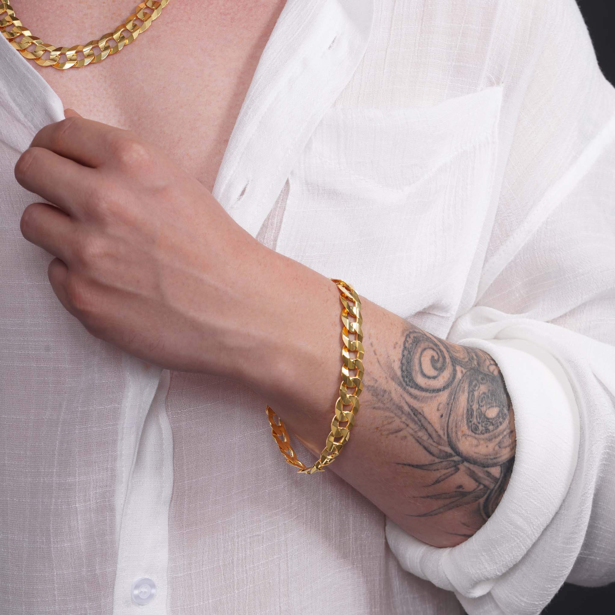 Men's solid gold curb bracelet worn on a hand with a tattoo, paired with a white shirt and gold chain necklace.