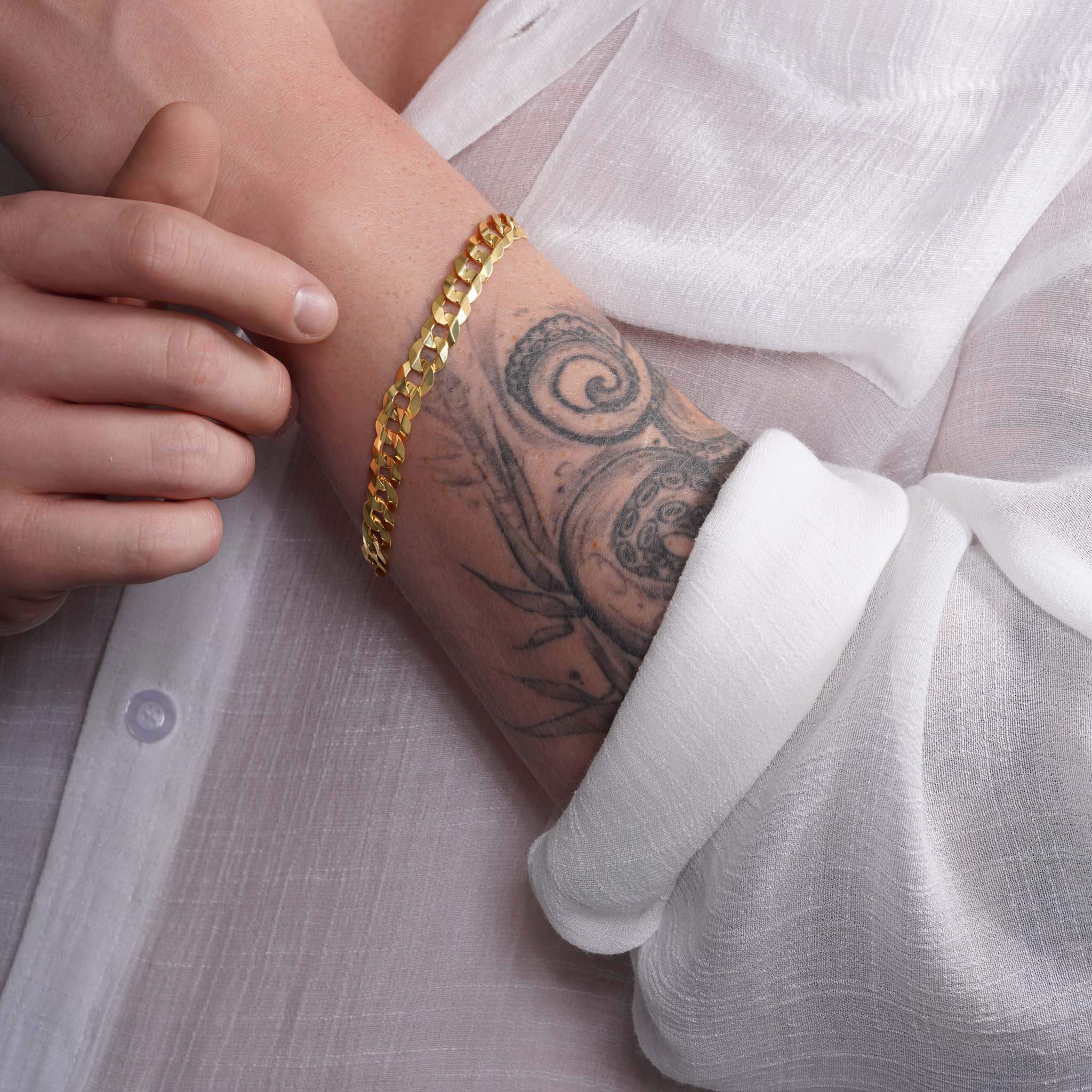 Close-up of a man's arm showcasing a 14K solid gold curb bracelet against a white shirt and tattoo.