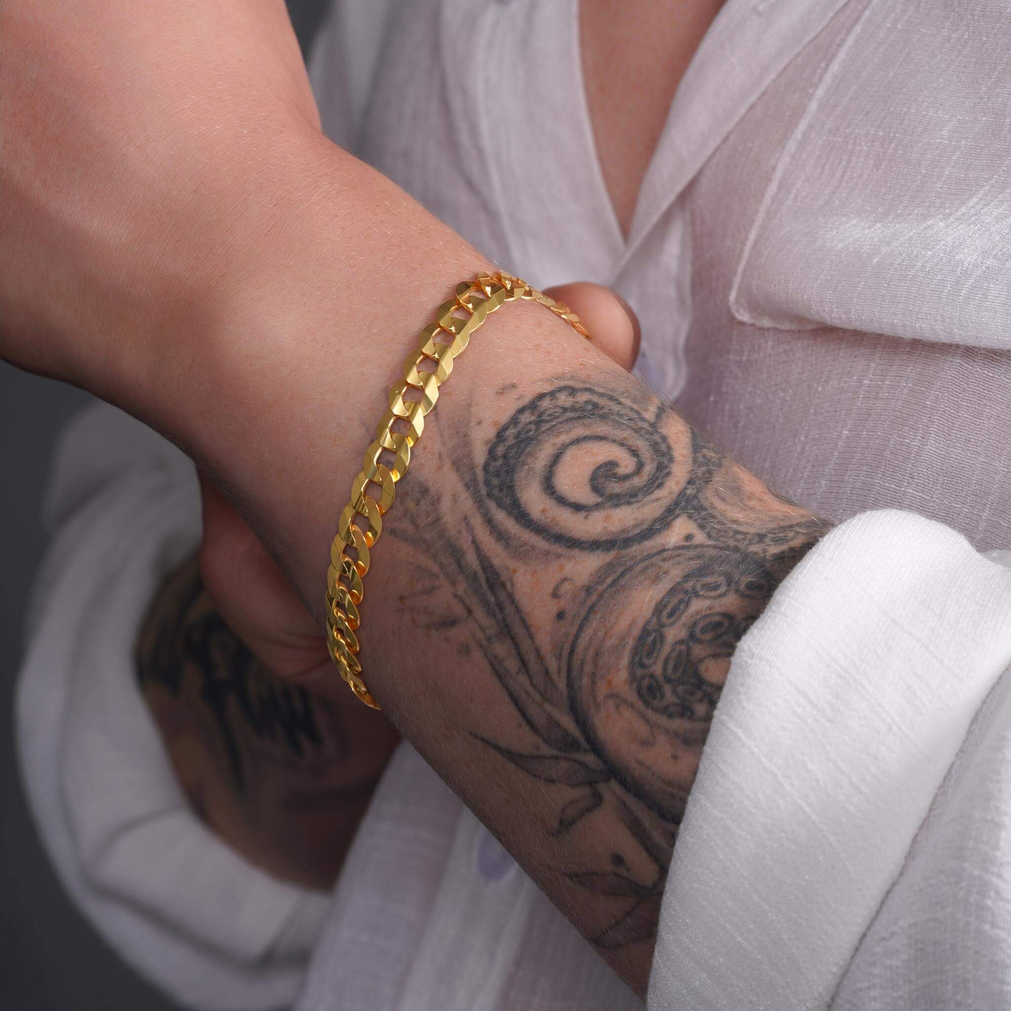 Close-up of a man's wrist wearing a 14K solid gold curb bracelet, showcasing a tattooed arm and elegant jewelry.