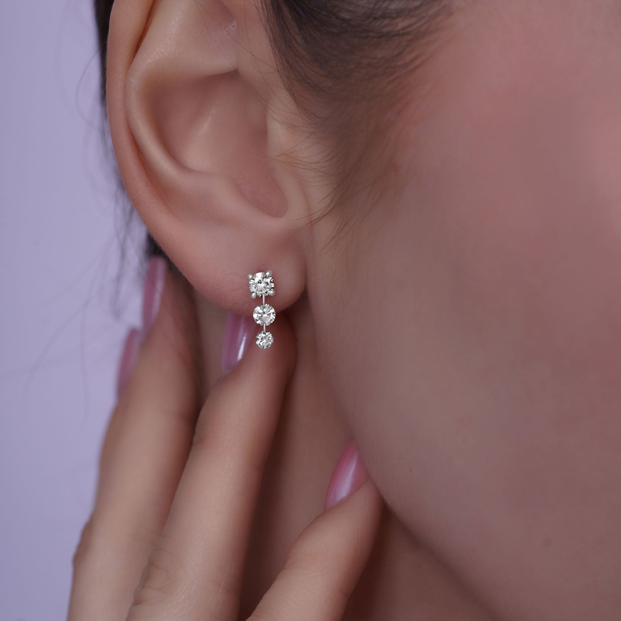 Close-up of a person wearing white gold stud earrings containing 3  dangling diamonds with a blurred background