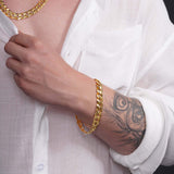 Men's solid gold curb bracelet worn on a hand with a tattoo, paired with a white shirt and gold chain necklace.
