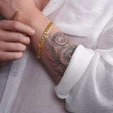Close-up of a man's arm showcasing a 14K solid gold curb bracelet against a white shirt and tattoo.