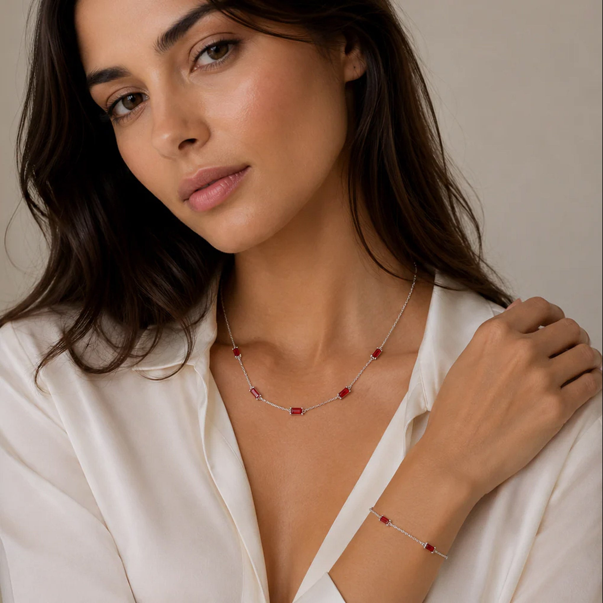 A woman wearing a delicate silver necklace and matching bracelet, each adorned with small red gemstone accents, against a soft beige background.