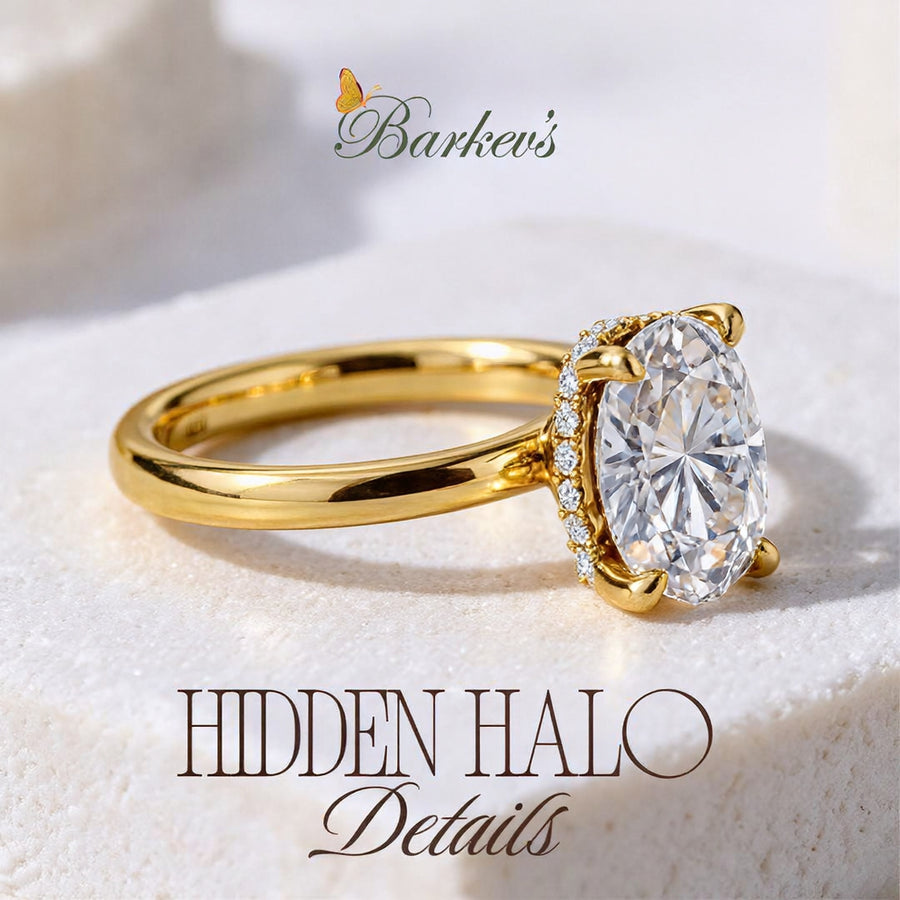 A gold ring with a large oval clear gemstone and small surrounding stones set around a hidden halo, shown on a soft white textured surface. The Barkev's logo appears above, and the text 'HIDDEN HALO Details' appears below.